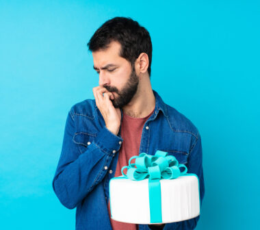 Young handsome man with a big cake over isolated blue background having doubts