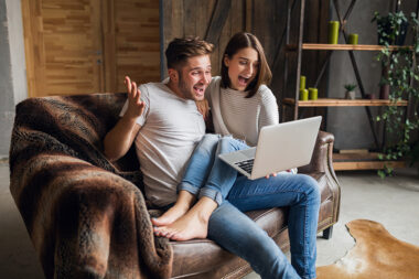 young smiling couple sitting on couch at home in casual outfit, love and romance, woman and man embracing, wearing jeans, spending relaxing time together, holding laptop, happy emotional