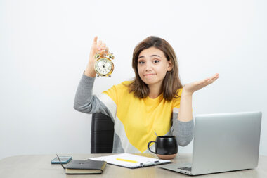 Photo of female office worker sitting at the desk and holding alarm clock